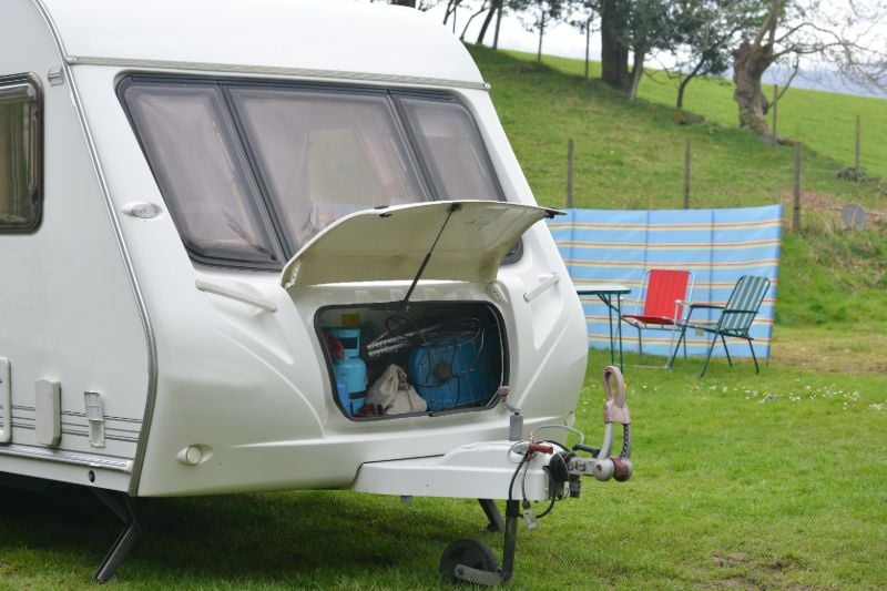 Close-up van de voorkant van een caravan met de opslagbox open op het platteland met ligstoelen en windscherm op de achtergrond
