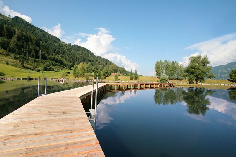 Houten steiger aan een rustig bergmeer bij Sportcamp Woferlgut, met groene heuvels en blauwe lucht op de achtergrond. 