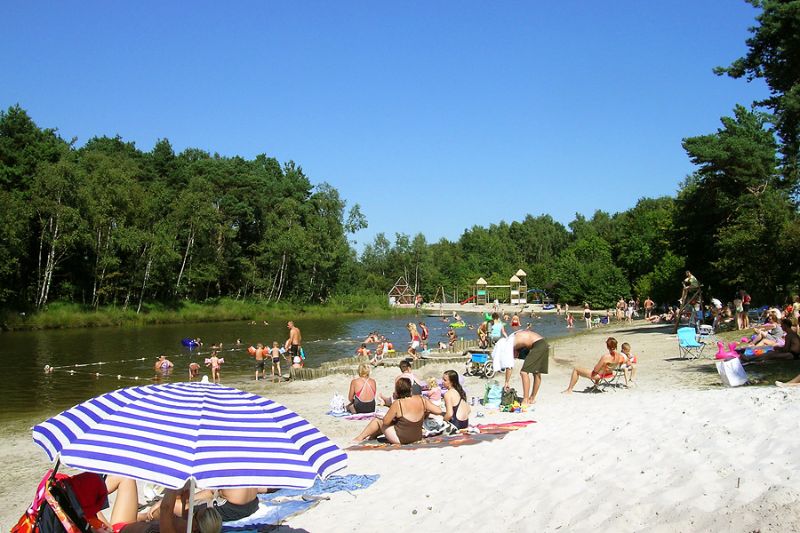 Zandstrand bij ArdenParks Baalse Hei met recreatieplas, spelende kinderen in het water en mensen die ontspannen in de zon aan de bosrand.