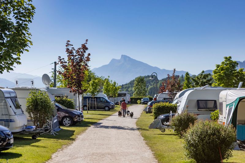 Kampeerplaats op Camp MondSeeLand met caravans en campers, met bergen op de achtergrond en een wandelaar met honden op het pad