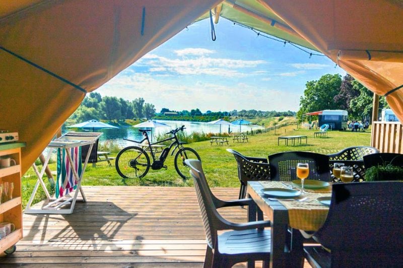 Uitzicht vanuit een safaritent op Camping De Boomgaard met terras, eettafel en fiets, gelegen aan een recreatieplas met ligweide en parasols.
