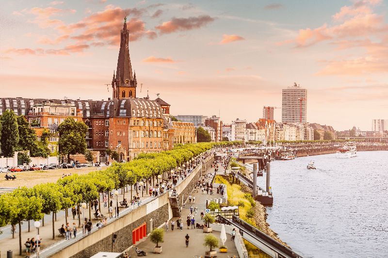 Skyline van Düsseldorf met de Rijnpromenade, wandelaars langs het water en historische gebouwen onder een warme avondlucht.
