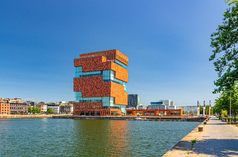 Het MAS Museum in Antwerpen aan het water, met het opvallende roodbruine gebouw en een promenade langs de kade onder een heldere blauwe lucht.