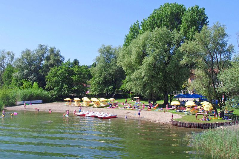 Strandje van Camping Village Lago Maggiore aan het Lago Maggiore met gele parasols en wit/rode waterfietsen. 